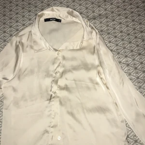 Beige sidenblus från Bik Bok - Elegant beige blus i siden från Bik Bok. Blusen har långa ärmar och knappar framtill, vilket ger en klassisk look. Perfekt för en stilren och sofistikerad outfit.