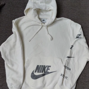 Vit hoodie från Nike - Säljer en stilren vit hoodie från Nike med silvrig logga på bröstet och text längs ärmen. Den har en klassisk känguruficka och justerbar huva med dragsko. Perfekt för en avslappnad look. 🖤 Lite skrynklig efter att ha legat längst ner i byrålådan 😊