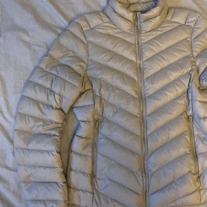 Beige dunjacka med dragkedja - Snygg beige dunjacka med dragkedja och hög krage. Jackan har en quiltad design och långa ärmar, perfekt för kyligare dagar. Den har praktiska fickor med dragkedjor på sidorna.