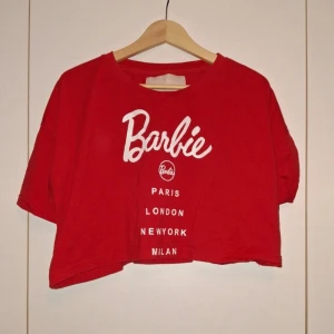 Röd Barbie t-shirt - Snygg röd Barbie t-shirt med tryck av städer som Paris, London, New York och Milan. T-shirten är kortärmad och har en croppad design, perfekt för en avslappnad stil.