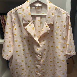 Rosa blommig skjorta från Monki - Säljer en söt rosa skjorta från Monki med ett blommigt mönster i gult och vitt. Skjortan har korta ärmar och knappar framtill. Perfekt för en somrig look! 🌼