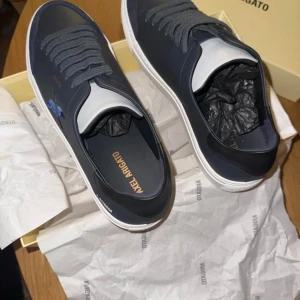 Navy sneakers från Axel Arigato - Snygga navy sneakers från Axel Arigato med en stilren design. Skorna har en broderad fågel på sidan och är tillverkade i skinn. De har en vit sula och matchande snörning för en modern look.