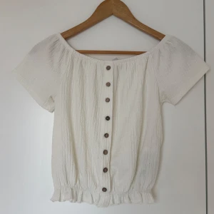 Vit offshoulder topp med knappar - Säljer en söt vit offshoulder topp med knappar framtill. Toppen har korta ärmar och en elastisk nederkant med volangdetalj. Perfekt för en somrig look!