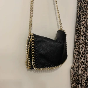 Svart axelväska med kedjedetaljer - Snygg svart axelväska från Tiamo i skinn med guldfärgade kedjedetaljer längs kanterna och som axelrem. Väskan har en praktisk storlek och stängs med magnetknapp. Perfekt för att ge en edgy touch till din outfit.