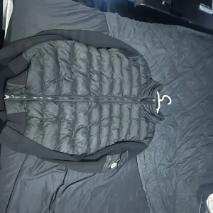 Svart dunväst från Moncler - Säljer en snygg svart dunväst från Moncler med dragkedja framtill. Västen har en quiltad design och en broderad logga på ärmen. Perfekt för kyligare dagar!