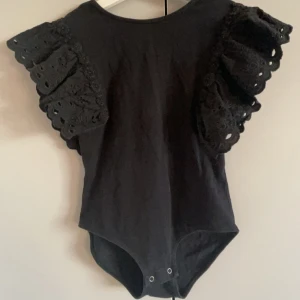 Svart body från Zara - Säljer en svart body från Zara med vackra spetsdetaljer på ärmarna. Den har en rund hals och knäppning i grenen. Perfekt för att ge en elegant touch till din outfit.
