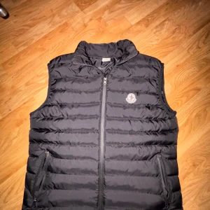 Svart dunväst från Moncler Storlek S - Snygg svart dunväst från Moncler med dragkedja och broderad logotyp på bröstet. Perfekt för kyligare dagar och kalla sommardagar.