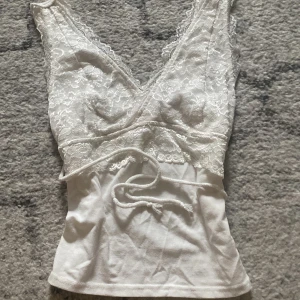 Vit spetslinne med omlott och knyt - Säljer ett sött vitt linne med spetsdetaljer och omlott-design. Toppen har en v-ringad fram och knytband i midjan. Perfekt för en somrig look!