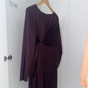 Mörkbrun långklänning - Elegant mörkbrun långklänning med en unik knutdetalj framtill. Klänningen har långa ärmar och är tillverkad i ett mjukt, glansigt material som ger en lyxig känsla. Perfekt för speciella tillfällen.