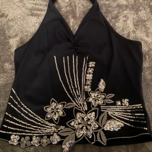 Svart halterneck topp med paljetter - Snygg svart halterneck topp med v-ringning och dekorativa paljetter i blommönster. Perfekt för en kväll ute eller festliga tillfällen. Toppen knyts i nacken och har en elegant design.