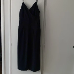 Svart byxdress med smala axelband - Elegant svart byxdress med smala axelband och omlott-design. Den har en v-ringad framdel och dragkedja i ryggen som är lite öppen. Perfekt för en stilren look.