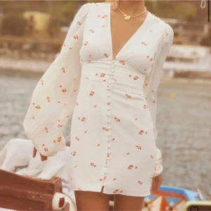  Vit klänning med blommönster från Adoore - Helt oanvänd och ny klänning!!Charmig vit klänning från Adoore med röda blommor. Den har en v-ringad design och långa puffärmar. Klänningen är knälång och har knappar framtill, vilket ger en elegant touch.❣️❣️kommer inte sälja för under 1500