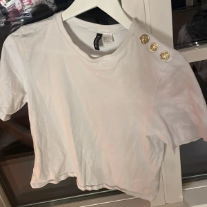 Vit kortärmad topp från H&M - Säljer en vit kortärmad topp från H&M med tre dekorativa guldknappar på axeln. Toppen har inga fläckar är i bra skick och har en rund halsringning. Perfekt för en stilren look.