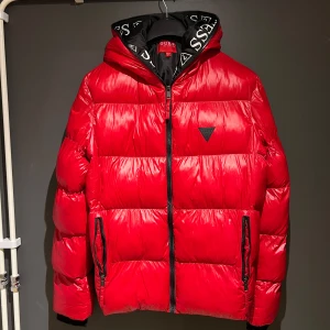 Röd pufferjacka från Guess - Snygg röd dunjacka från Guess med huva och dragkedja. Jackan har en quiltad design och svarta detaljer på dragkedjorna. Perfekt för kyliga dagar. Står L på den men jag är 183 och inte så stor och passa mig perfekt! Passar inte priset så kom med förslag👑