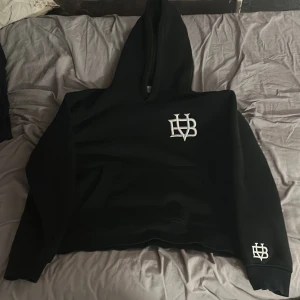Svart hoodie från Bvaldi - Säljer en svart hoodie från Bvaldi med en broderad vit logga på bröstet och ärmen. Den har en klassisk passform med huva och långa ärmar. Perfekt för en avslappnad stil. Skulle säga att den passar också storleken M