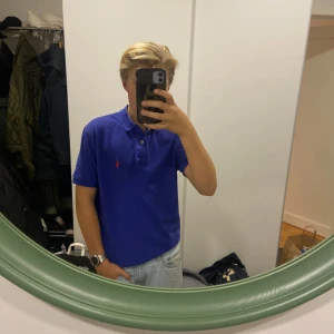 Blå pikétröja från Ralph Lauren - Säljer en klassisk blå pikétröja från Ralph Lauren med röd logga på bröstet. Tröjan har korta ärmar och knappar vid kragen. Perfekt för en stilren look.