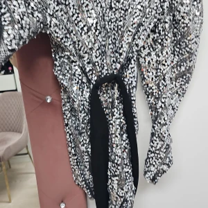 Glittrig klänning med knytband - Snygg glittrig klänning med ett svart knytband i midjan. Perfekt för en festlig kväll med sitt iögonfallande mönster och glittriga detaljer.