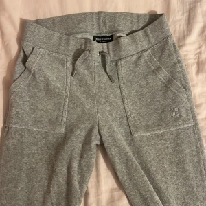 Grå mjukisbyxor från Juicy Couture - Grå mjukisbyxor från Juicy Couture med snörning i midjan och broderad logga på framsidan. Byxorna har praktiska fickor både fram och bak med knappdetaljer. Perfekta för en avslappnad stil.