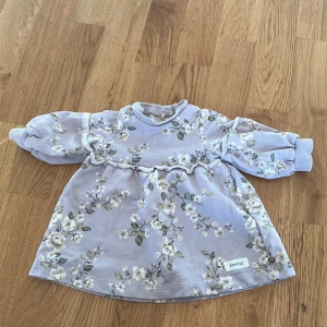 Blommig klänning från Newbie - Söt blommig klänning från Newbie i storlek 56. Klänningen har ett vackert blommönster i ljusa toner och långa ärmar med ribbade muddar. Perfekt för små barn som vill vara både bekväma och stiliga.