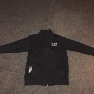 Svart jacka från Emporio Armani EA7 - Snygg svart jacka från Emporio Armani EA7 med dragkedja och vita detaljer. Perfekt för en stilren look. Jackan har långa ärmar och en hög krage.