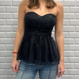 Svart glittrig tubtopp - Snygg svart tubtopp med glittriga detaljer. Toppen är axelbandslös och har en lätt utsvängd nederdel som ger en fin siluett. Perfekt för en kväll ute eller festliga tillfällen.