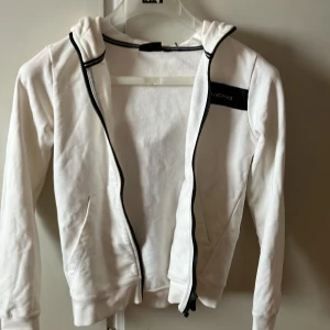 Vit hoodie från Sail Racing - Säljer en snygg vit hoodie från Sail Racing i storlek 150. Den har en dragkedja framtill och en cool logga på huvan. Perfekt för både vardag och chill. Hoodien är i bra skick och redo för nya äventyr!