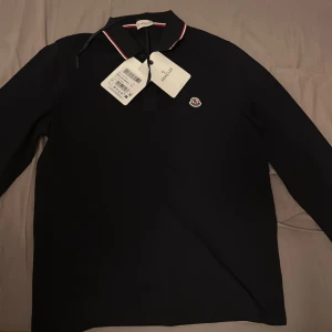 Svart pikétröja från Moncler - Säljer en stilren svart pikétröja från Moncler med lång ärm och klassisk krage. Tröjan har en broderad Moncler-logga på bröstet och detaljer i rött och vitt på kragen. Perfekt för en elegant och avslappnad look.