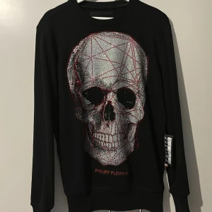 Svart tröja med dödskalle från Philipp Plein - Philipp Plein med en stor, detaljerad dödskalle i grått och rött på framsidan. Tröjan har långa ärmar och är i mycket bra skick. Köpt på Miinto för ca 3 år sedan 