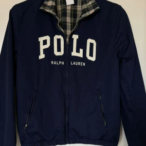 Mörkblå jacka från Ralph Lauren - Säljer en snygg mörkblå jacka från Ralph Lauren med stort POLO-tryck på framsidan. Jackan har en dragkedja och två fickor. Insidan är rutig i grönt och vitt, vilket ger en cool kontrast. Perfekt för höst och vår! Storlek 18-20 år barn