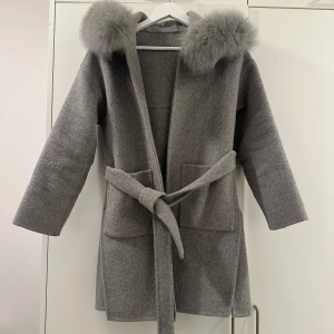 Grå kappa med pälsdetaljer - Elegant grå kappa med stor huva dekorerad med mjuk päls. Kappan har ett knytbälte i midjan och långa ärmar. Perfekt för kyliga dagar när du vill hålla dig varm och stilren.