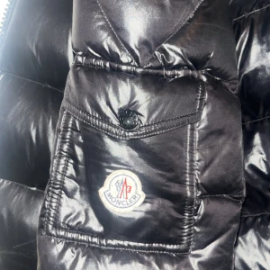 Svart pufferjacka från Moncler - Snygg svart pufferjacka från Moncler med glansig finish. Jackan har en dragkedja framtill och en praktisk ficka med knappdetalj. Perfekt för kyliga dagar med stil. Hör av dig för fler bilder!