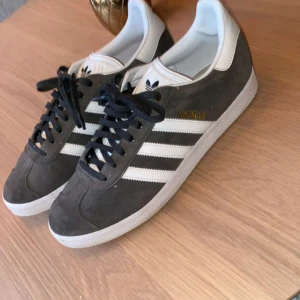 Adidas Gazelle sneakers i grått och vitt - Säljer ett par klassiska Adidas Gazelle sneakers i grå mocka med vita detaljer och snörning. Skorna har den ikoniska tre-randiga designen och en vit sula. Perfekta för en stilren och sportig look.