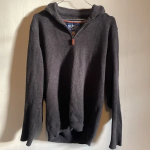 Mörkgrå tröja från Superdry - Säljer en snygg mörkgrå tröja från Superdry i bra skick. Den har en dragkedja vid halsen och är perfekt för höst och vinter. Tröjan är långärmad och har en bekväm passform. Perfekt för både vardag och lite mer avslappnade tillfällen.