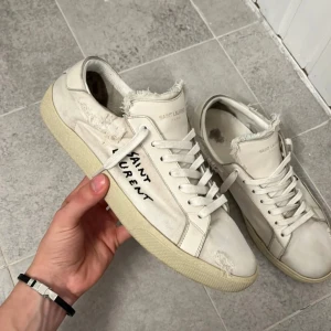 Vita sneakers från Saint Laurent - Säljer ett par vita sneakers från Saint Laurent i bra skick. De har en cool, sliten look med fransiga detaljer och handskriven logga på sidan. Perfekta för en avslappnad stil med en touch av lyx. Skorna kommer med originalförpackning.