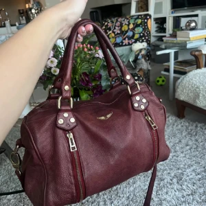 Vinröd handväska från Zadig & Voltaire - Snygg vinröd skinnväska i skinn från Zadig & Voltaire. Väskan har två handtag och detaljer i guld, samt praktiska dragkedjor på sidorna. Saknar en liten guld platta på höger sida. Men går att köpa till.  Perfekt för både vardag och fest! Köpt begagnad på Vestaire. 