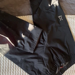 Svarta regnbyxor från Haglöfs - Säljer ett par svarta regnbyxor från Haglöfs i bra skick. Perfekta för regniga dagar med elastisk midja för bekväm passform. Byxorna är tillverkade i ett slitstarkt syntetmaterial som håller dig torr.  (Damm och solen gör att de vita prickarna syns på bild, ignorera det). Helt nya, använda 1-2 gånger)