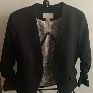 Svart kavaj från H&M - Elegant svart kavaj från H&M med blommigt foder. Kavajen har en stilren design utan krage och långa ärmar. Perfekt för en chic och sofistikerad look.