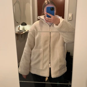 Vit teddyjacka med dragkedja - Mysig vit teddyjacka med dragkedja. Perfekt för kyliga dagar med sin fluffiga och varma design. Jackan har en avslappnad passform och stora fickor framtill.