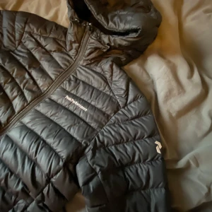 Svart dunjacka från Peak Performance - Säljer en svart dunjacka från Peak Performance i bra skick. Jackan har en quiltad design med dragkedja och huva, perfekt för kyliga dagar. Den är långärmad och passar bra för både höst och vinter. Perfekt för dig som vill hålla stilen och värmen samtidigt! 