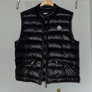 Svart dunväst från Moncler  - Säljer en snygg svart dunväst från Moncler i mycket bra skick. Västen har en glansig finish och är perfekt för användning året om. Den har en dragkedja framtill och två praktiska fickor med dragkedja. Moncler-loggan är broderad på bröstet. Modell ”Gui” storlek M.