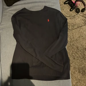 Svart tröja från Ralph Lauren - Säljer en stilren svart tröja från Ralph Lauren med det klassiska röda logotypbroderiet på bröstet. Tröjan har långa ärmar och är perfekt för både vardag och lite finare tillfällen. Den är i bra skick och passar perfekt till höst- och vårgarderoben.