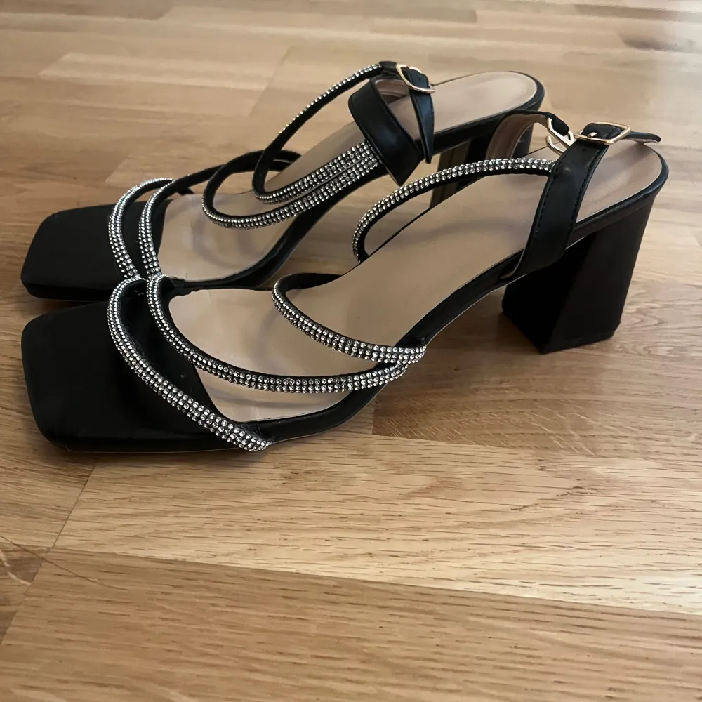Eleganta svarta klackar med fyrkantig tå och klack. Klackarna har flera remmar dekorerade med glittrande strass och justerbara spännen för perfekt passform.. Kengät.