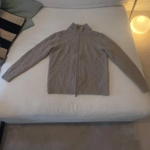  Massimo Dutti cardigan - Säljer en stilren beige stickad kofta från Massimo Dutti. Koftan har en dragkedja framtill och är långärmad med ribbade detaljer vid ärmslut och nederkant. Perfekt för kyligare dagar. Huvan har dock tappats bort, men det går att köpa en ny och tillsätta vid kragen . Därav priset 