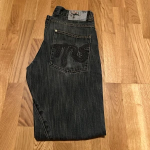 TRS Mörkblå jeansbyxor med broderad bakficka - Snygga mörkblå jeans från TRS med en unik broderad design på bakfickan. Byxorna har en klassisk femficksmodell och en rak passform. Perfekta för en avslappnad stil.