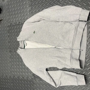 Grå hoodie från Lacoste - Säljer en stilren grå hoodie från Lacoste i mycket bra skick. Den har en dragkedja framtill och det klassiska krokodilmärket på bröstet. Perfekt för både vardag och avslappnade tillfällen. Passformen är normal och materialet känns mjukt och bekvämt. Missa inte chansen att äga denna snygga tröja! 🐊