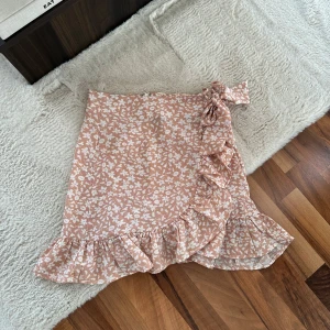 Rosa blommig kjol  - Söt blommig omlottkjol i rosa med volangdetaljer. Kjolen har en knytning i sidan för att justera passformen. Perfekt inför sommaren