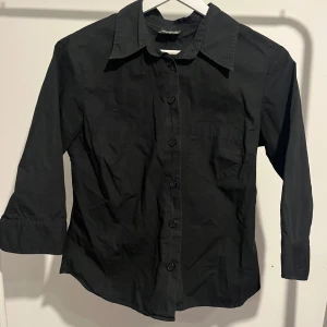 Svart skjorta med trekvartsärm - Snygg svart skjorta med trekvartsärm och knappar framtill. Perfekt för en stilren look. Skjortan har en klassisk krage och en enkel bröstficka.