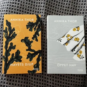 Havets djup och Öppet hav av Annika Thor - Två böcker av Annika Thor: 'Havets djup' med en orange omslag prydd med svarta korallmotiv och 'Öppet hav' med ett grått omslag dekorerat med en vit sjal med gula blommor. Perfekt för den som älskar spännande och känslosamma berättelser.