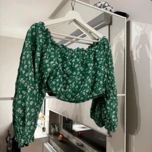 Grön blommig offshoulder-top - Snygg grön offshoulder-top med vita blommor. Toppen har långa ärmar och en luftig passform, perfekt för vår och sommar.