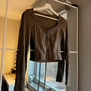 Brun långärmad topp med knyt - Snygg brun långärmad topp med knyt framtill. Toppen har en v-ringad design och är croppad, vilket ger en trendig look. Perfekt för att styla med jeans eller kjol.